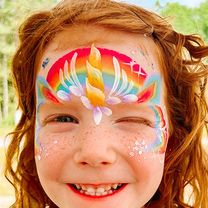 A smiling little redheaded girl with a unicorn face paint design with an orange horn, and painted sparkels with sparkly glitter on her cheeks.