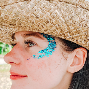 Woman turned to the side wearing a straw hat with a blue glitter wave design around her left eye at her temples.